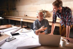 Two men going over shed plans on paper and a laptop in a workshop